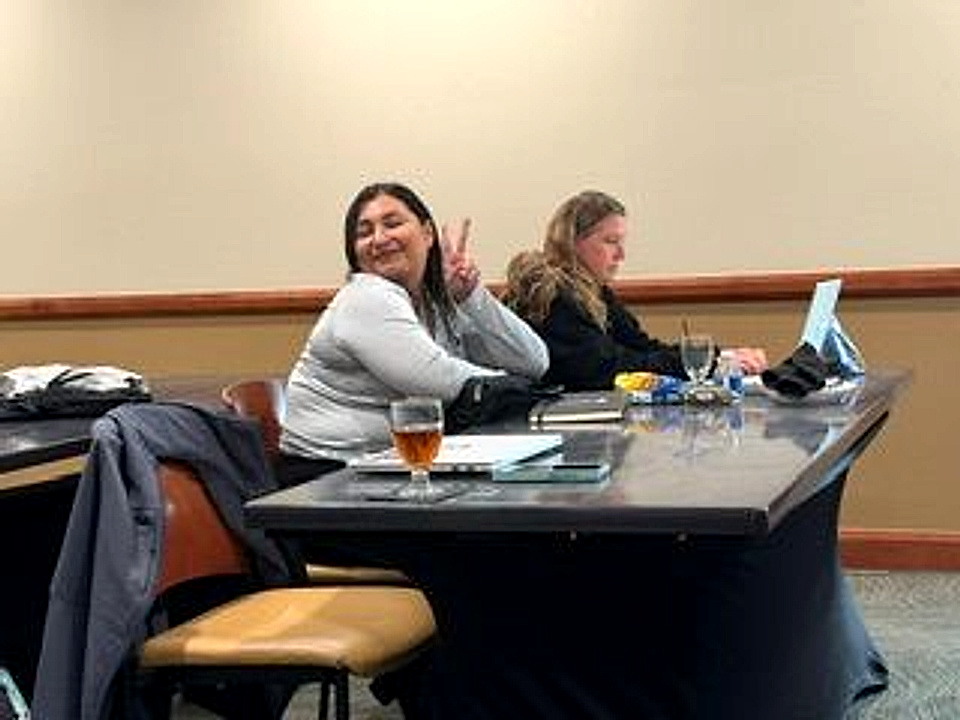 Two workshop participants at a table with an open laptop and notebooks; one turns toward the camera smiling and holding up a peace sign